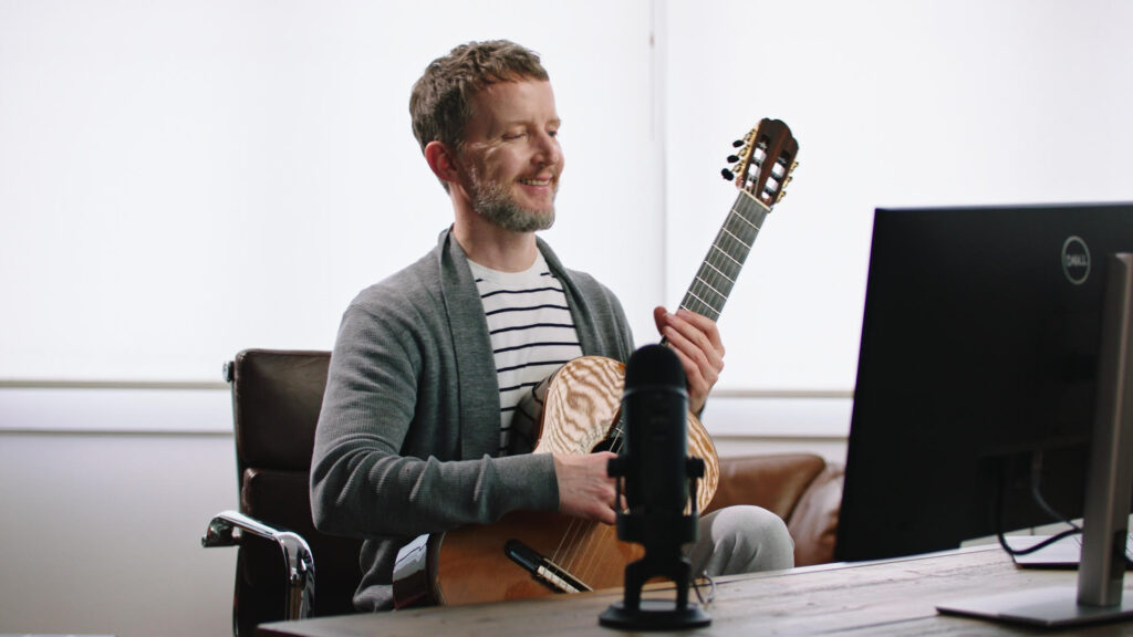 Martin Fogel holding a classical guitar in front of mic and screen during an online lesson at the Fogel Academy.